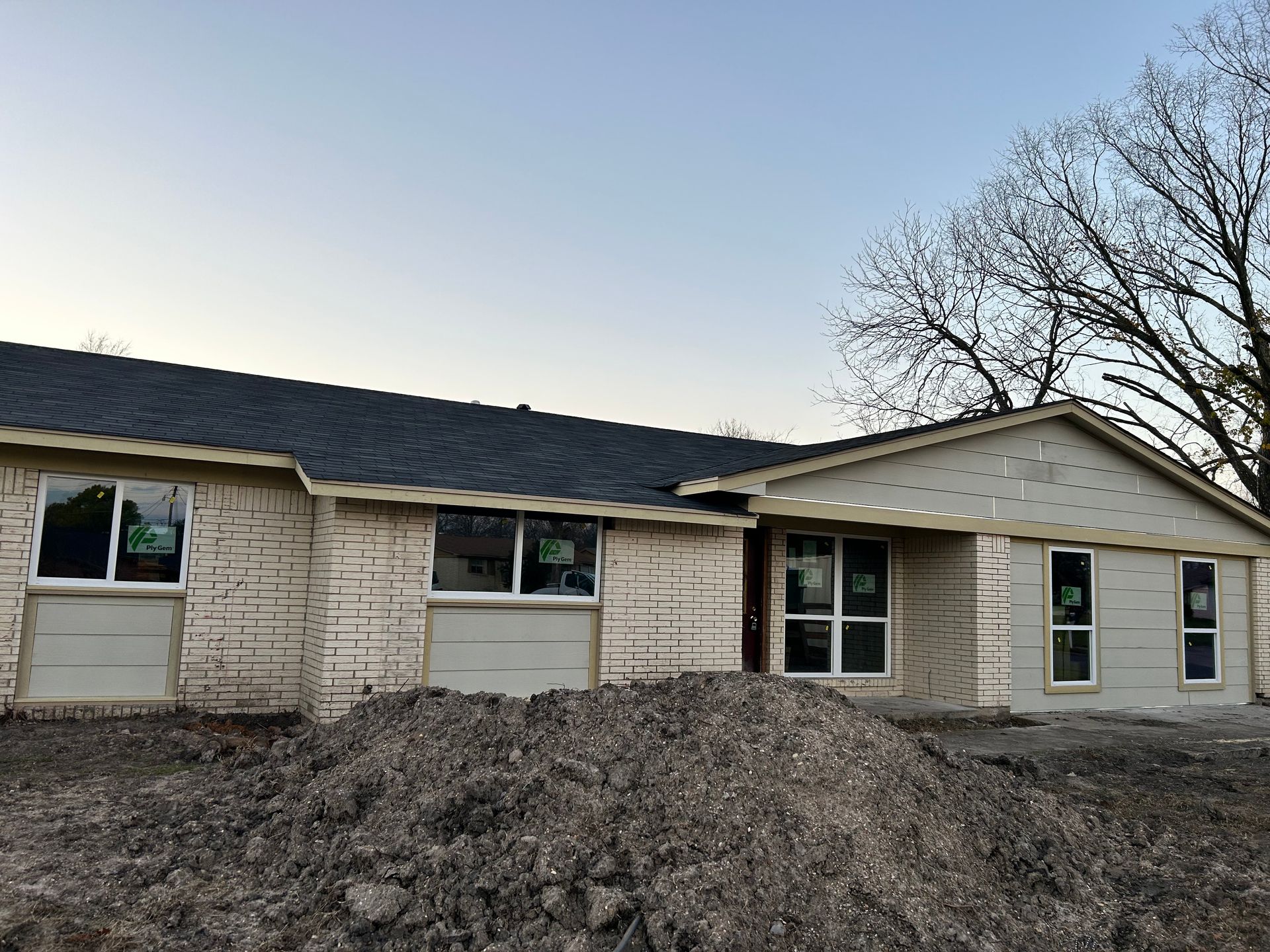 New construction house, light brick siding, windows, dark roof, dirt pile in the foreground, sky in the background.