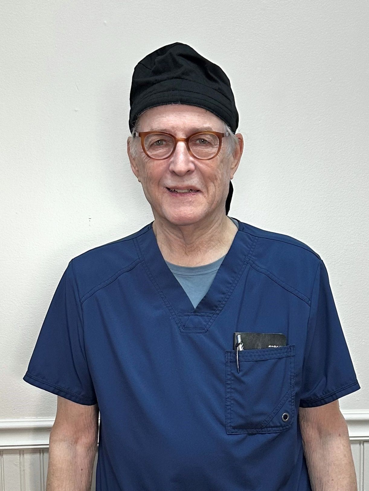 Man in blue scrubs, glasses, and a black cap, smiling against a white wall.