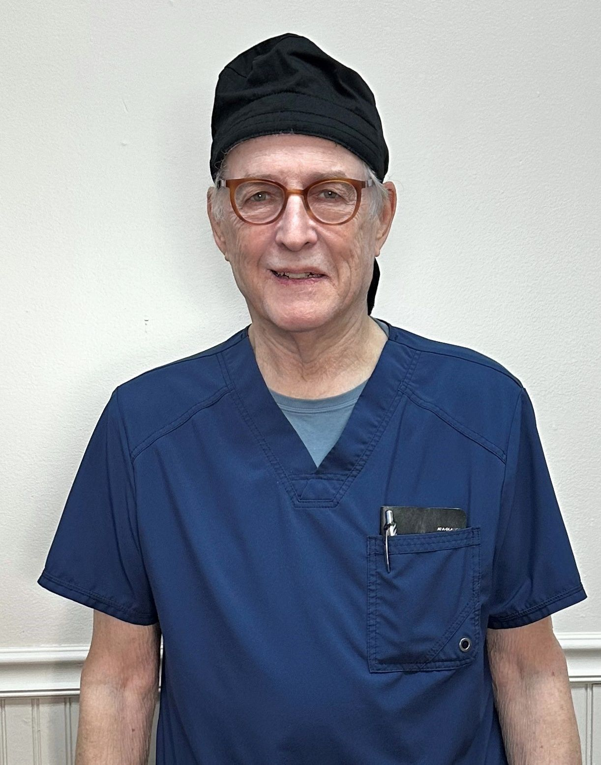 Person in blue scrubs and black cap standing against a plain white wall, facing the camera.