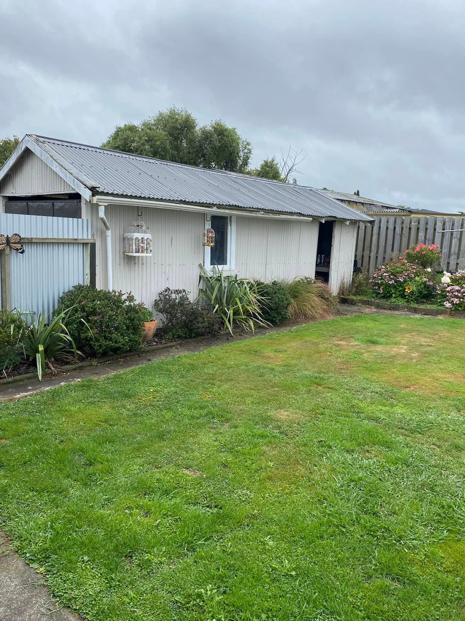 A small, weathered shed with a gray roof and a garden in front. Overcast sky.