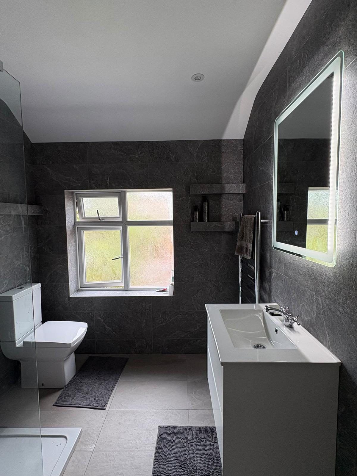 Walk-in shower room with grey slate-effect tiles, a white vanity unit and a backlit mirror, fitted by our team