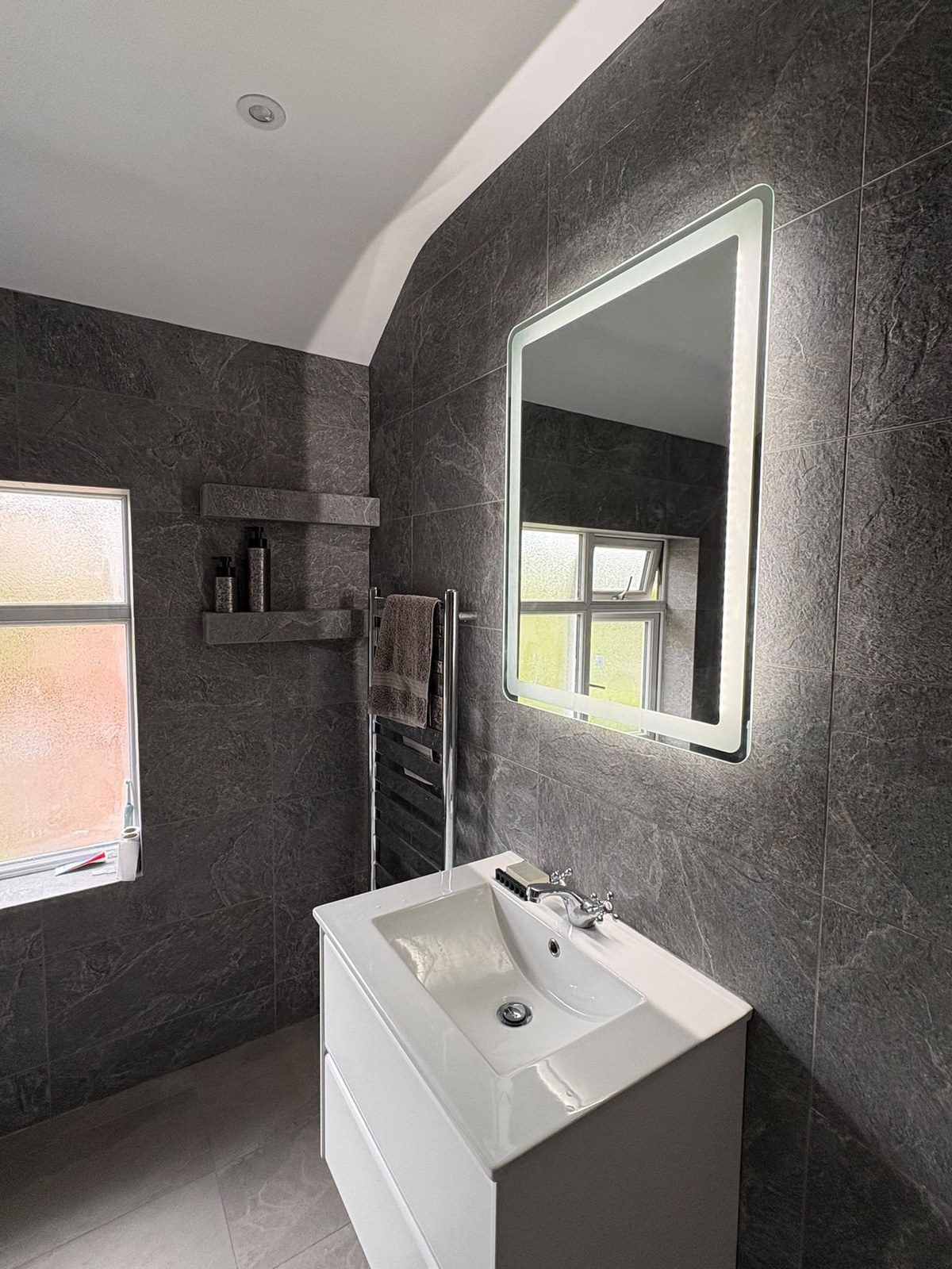 White two-drawer vanity unit under a backlit mirror, with tiled shelves beside the window