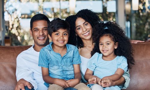 Family of four smiling on a couch, with two children in blue shirts seated between their parents.