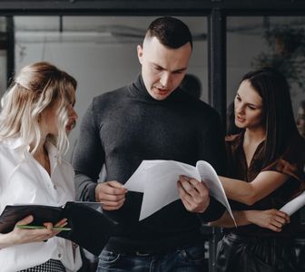 Three coworkers review printed documents in a modern office, standing closely and discussing them.