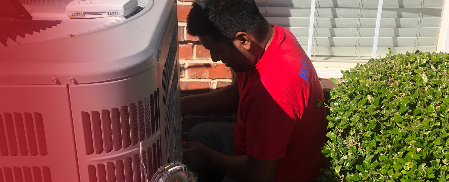 A man in a red shirt is working on an air conditioner outside of a house.
