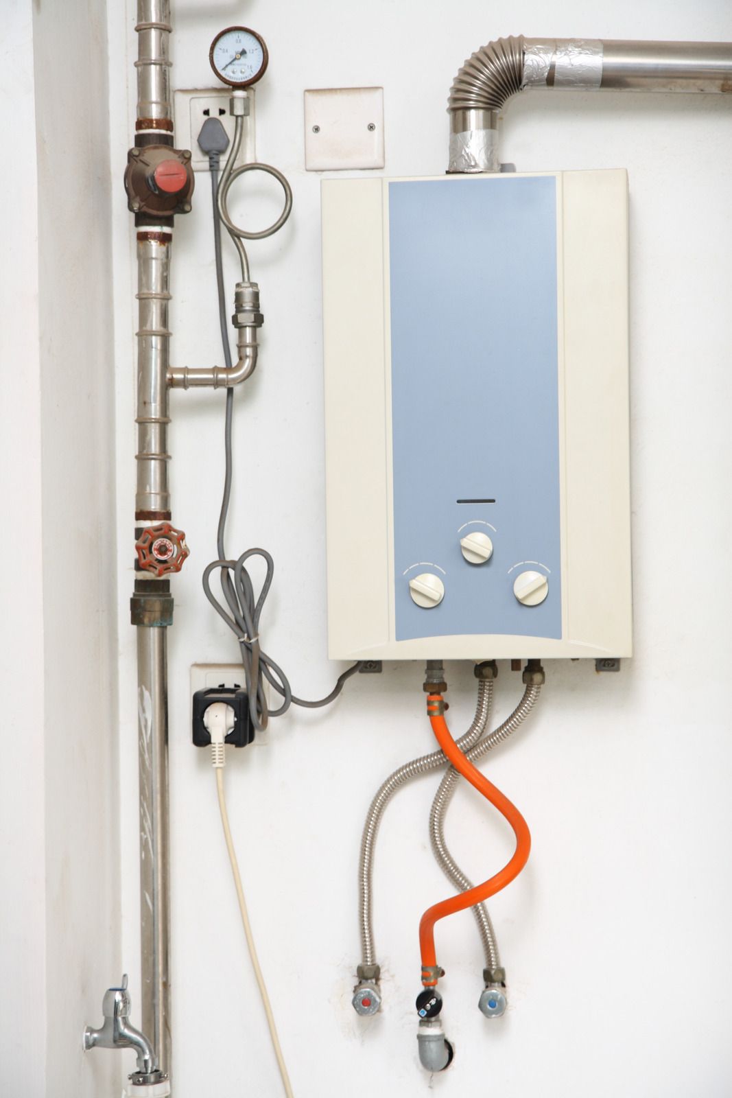 White water heater mounted on a wall with pipes, gauges, and electrical cord.