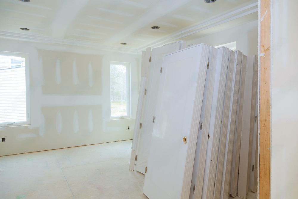 Bright unfinished room with drywall, white walls, windows, and stacked doors leaning on the right