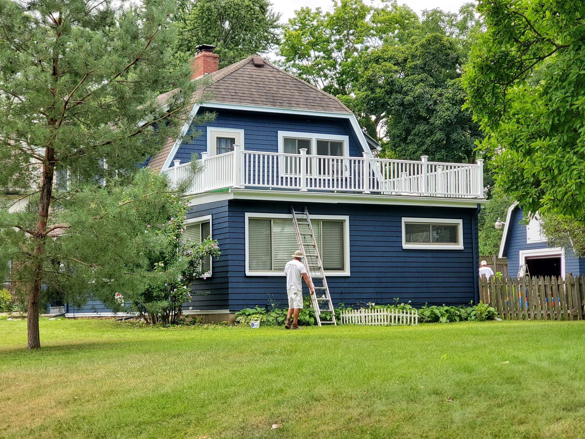 Blue house with a white balcony. Person on a ladder. Green lawn.