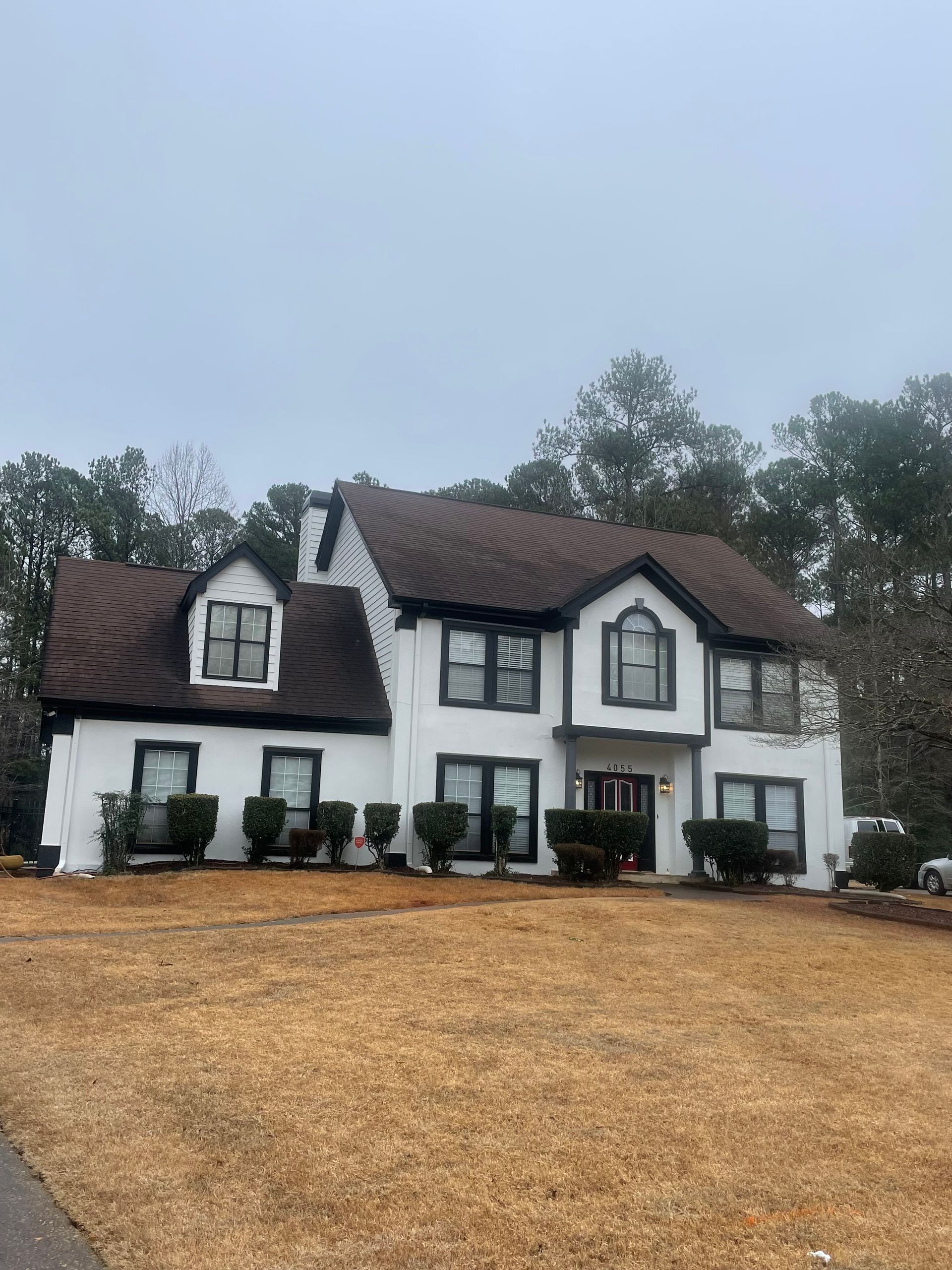 White two-story house with black trim and a brown roof, sitting on a brown lawn under a cloudy sky.