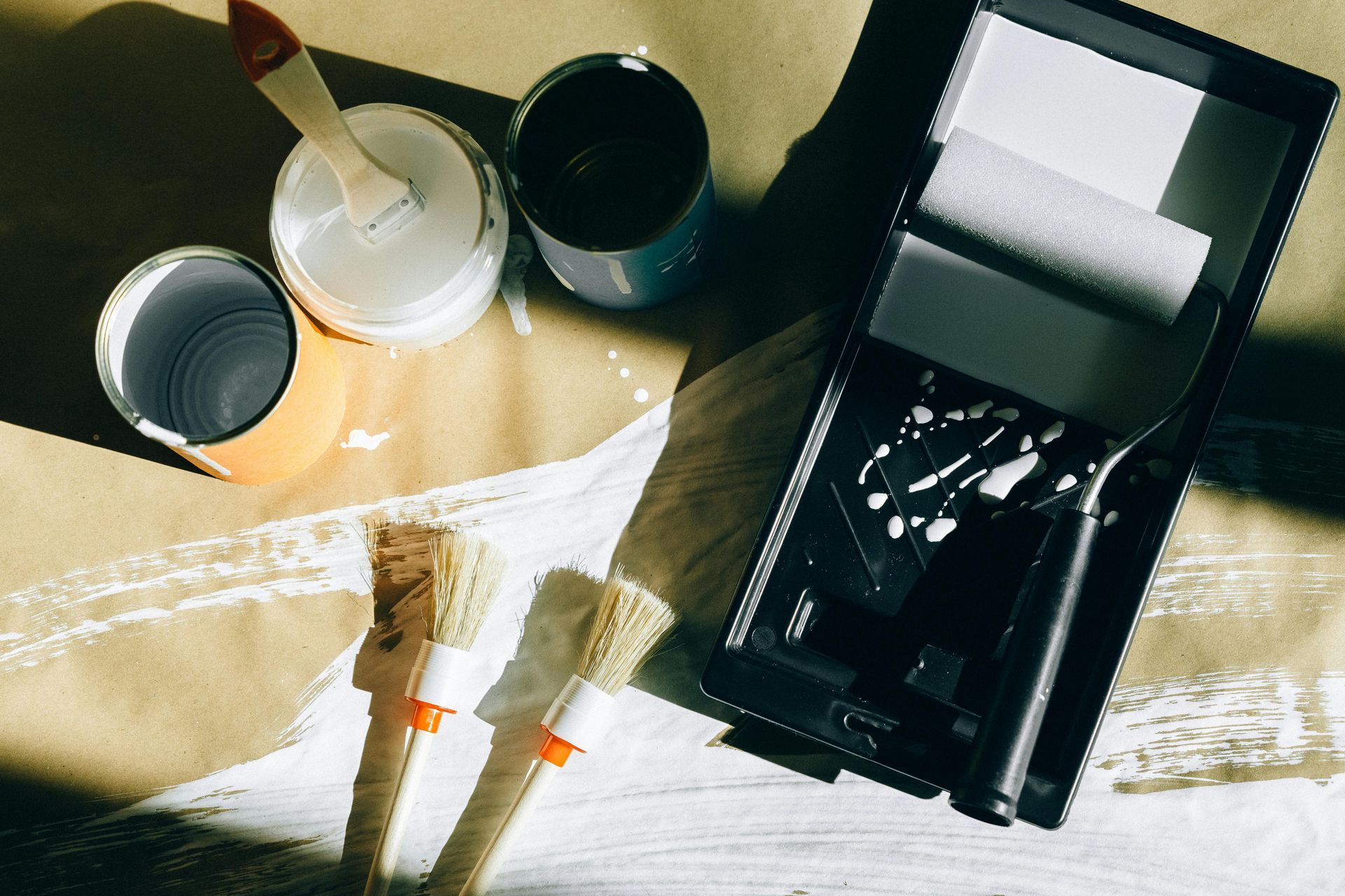 Paint cans, brushes, and roller tray with white paint on brown paper.