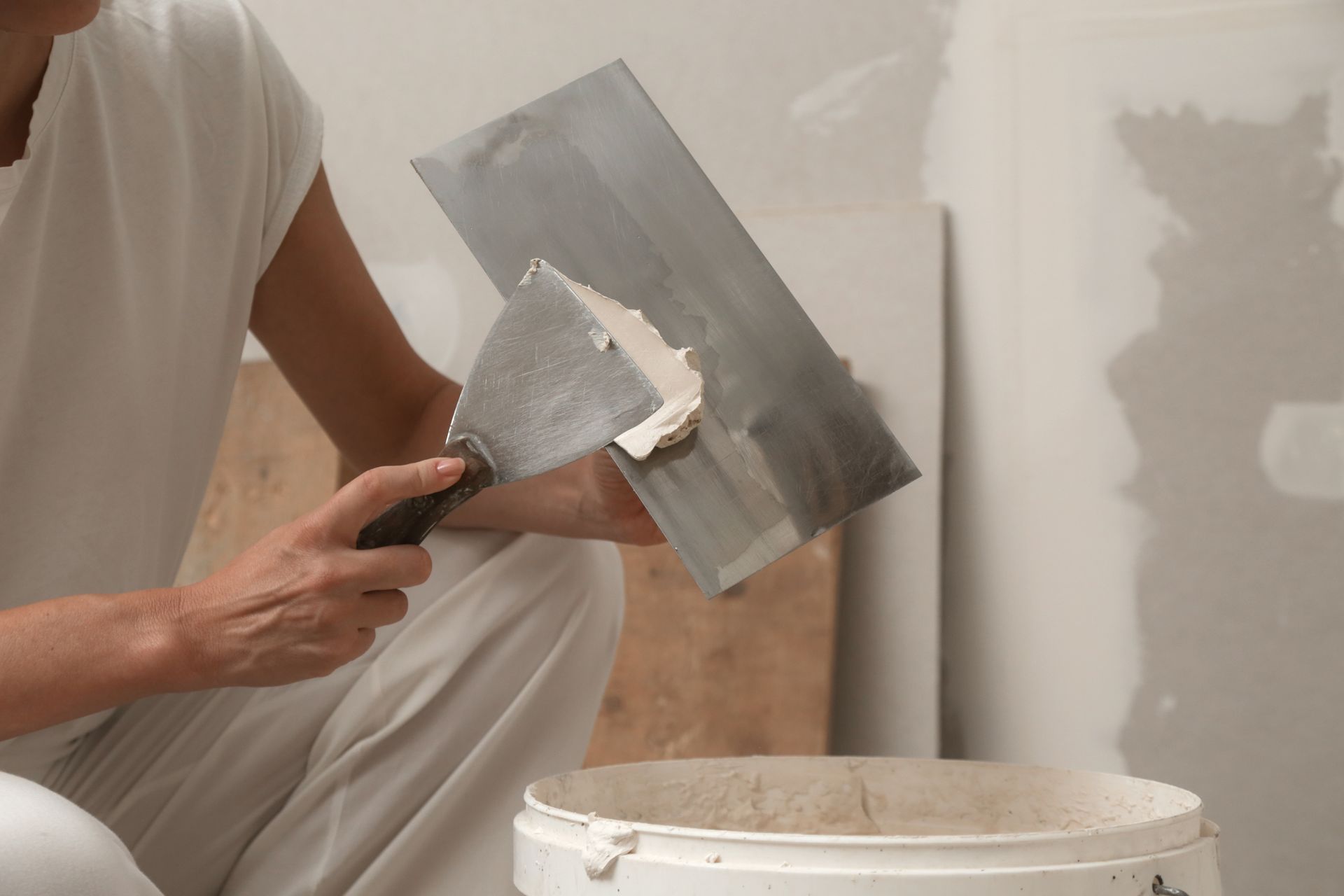 Person using a putty knife to apply joint compound to a wall, working from a bucket.