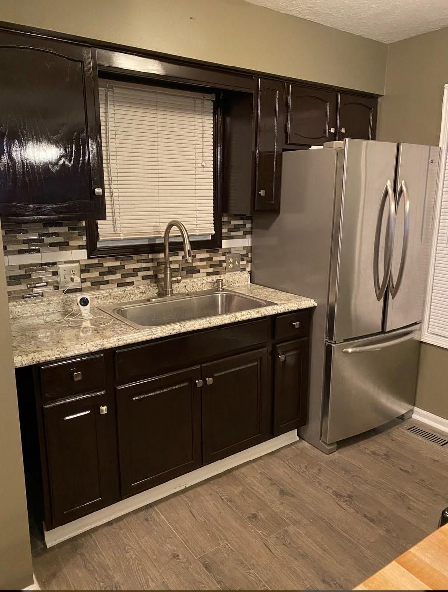 Dark brown kitchen cabinets, stainless steel refrigerator, sink under a window, and wood-look flooring.