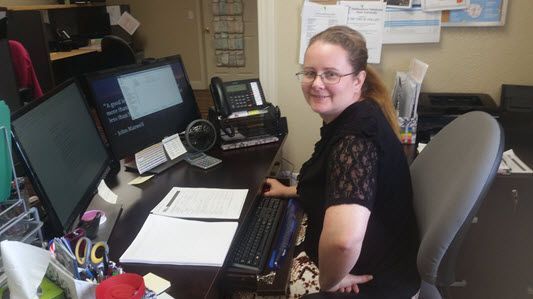 A woman is sitting at a desk with two computer monitors and a phone.