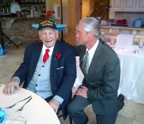 Two men are sitting at a table with one wearing a hat that says veteran