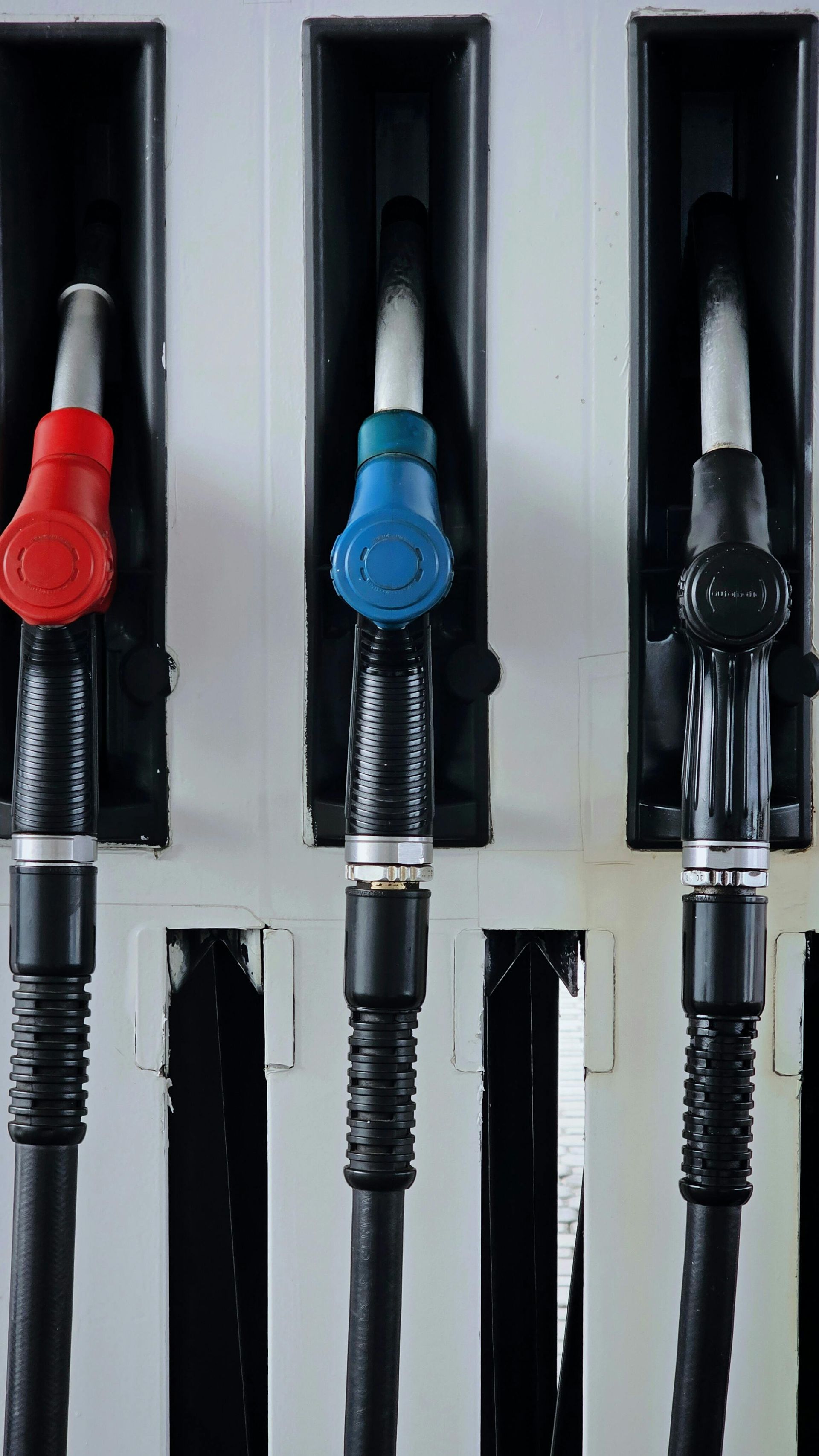 Three gas pump nozzles with red, blue, and black handles, hanging on a white pump station.