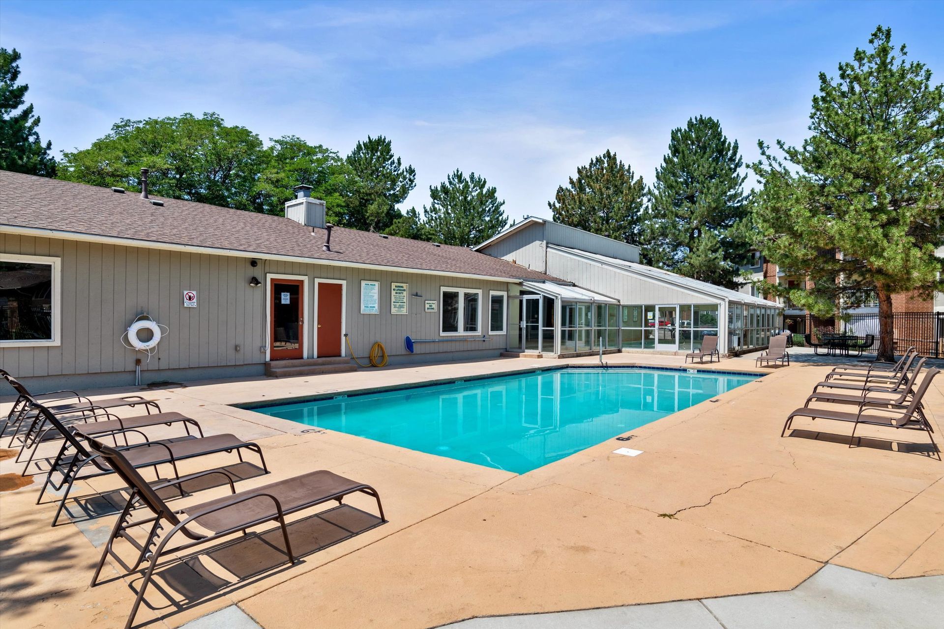 Poolside view: blue pool, lounge chairs, beige building, trees, and bright blue sky.