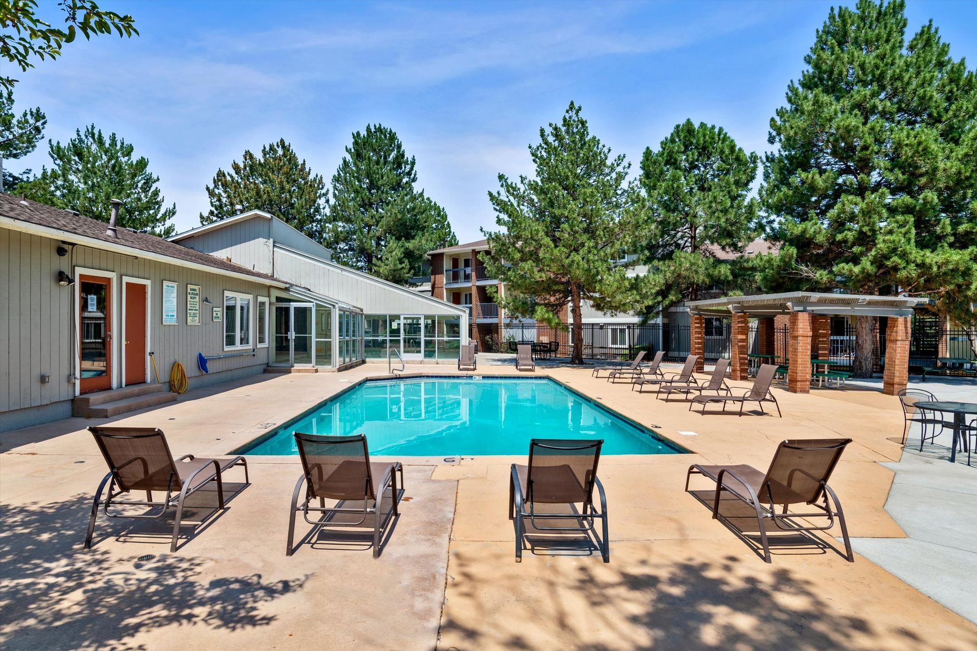 Swimming pool surrounded by lounge chairs, buildings, and trees under a blue sky.