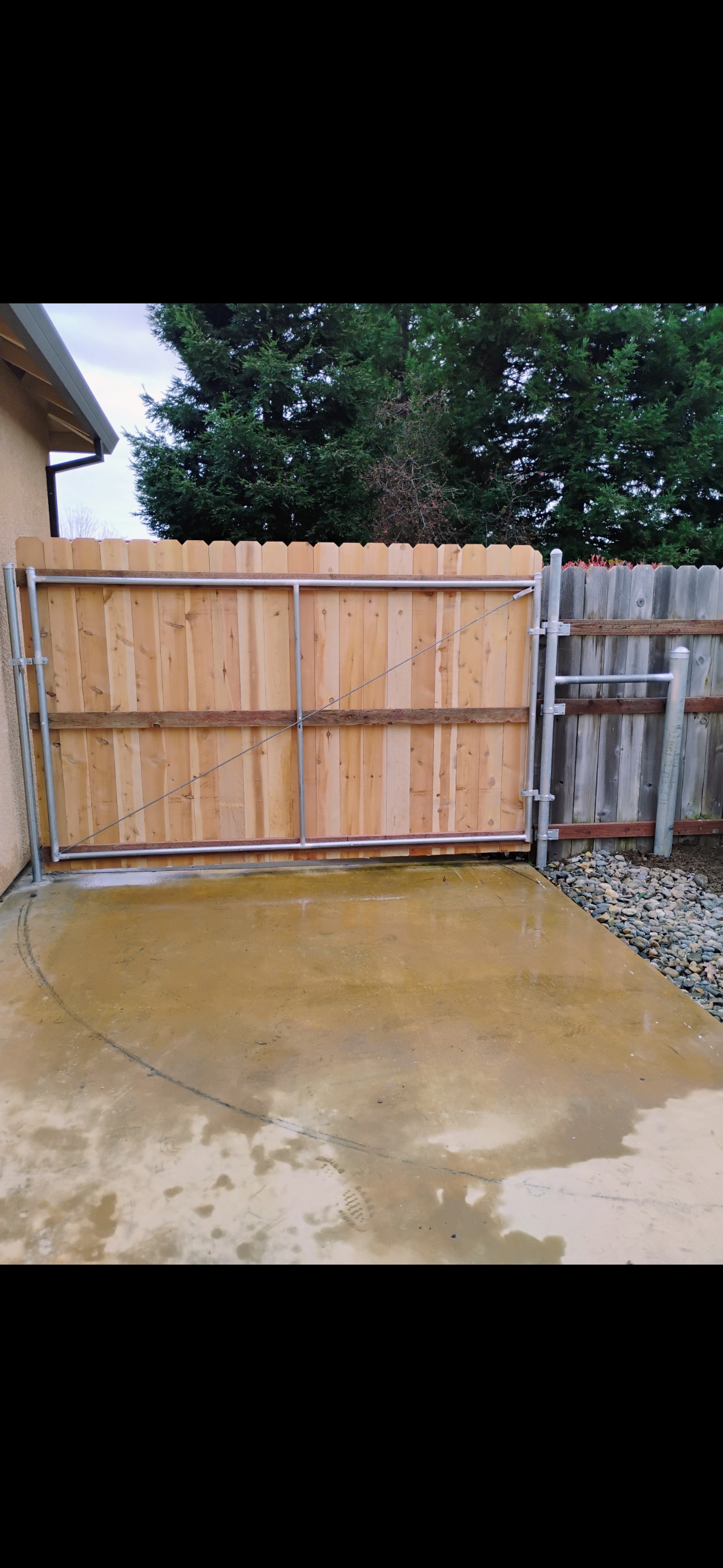 Wooden fence gate on a concrete patio. Trees in the background. Gray and brown tones. Wooden fence and gate with concrete surface, trees visible in the background.