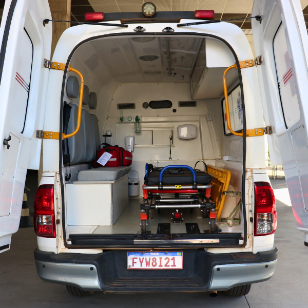 Ambulância com a traseira aberta, contendo maca e equipamentos médicos; van branca estacionada ao ar livre.