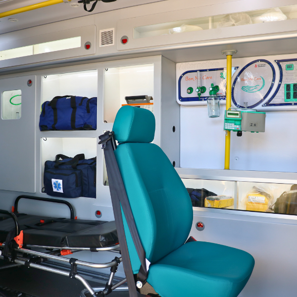 Interior de uma ambulância com uma cadeira médica verde-azulada, painéis de equipamentos e compartimentos de armazenamento.