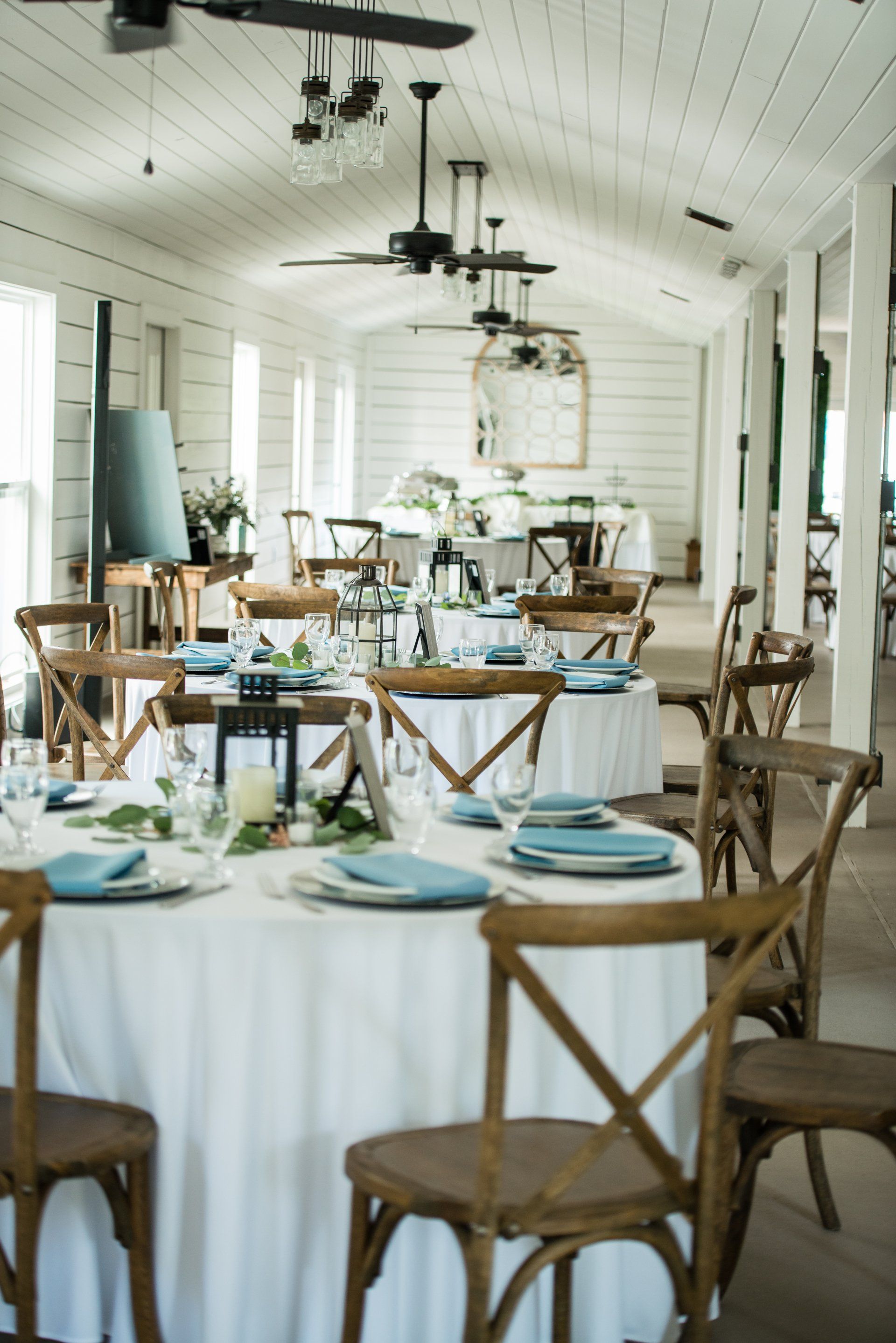 Bright white event dining room with round tables, blue napkins, and wooden chairs under black ceiling fans