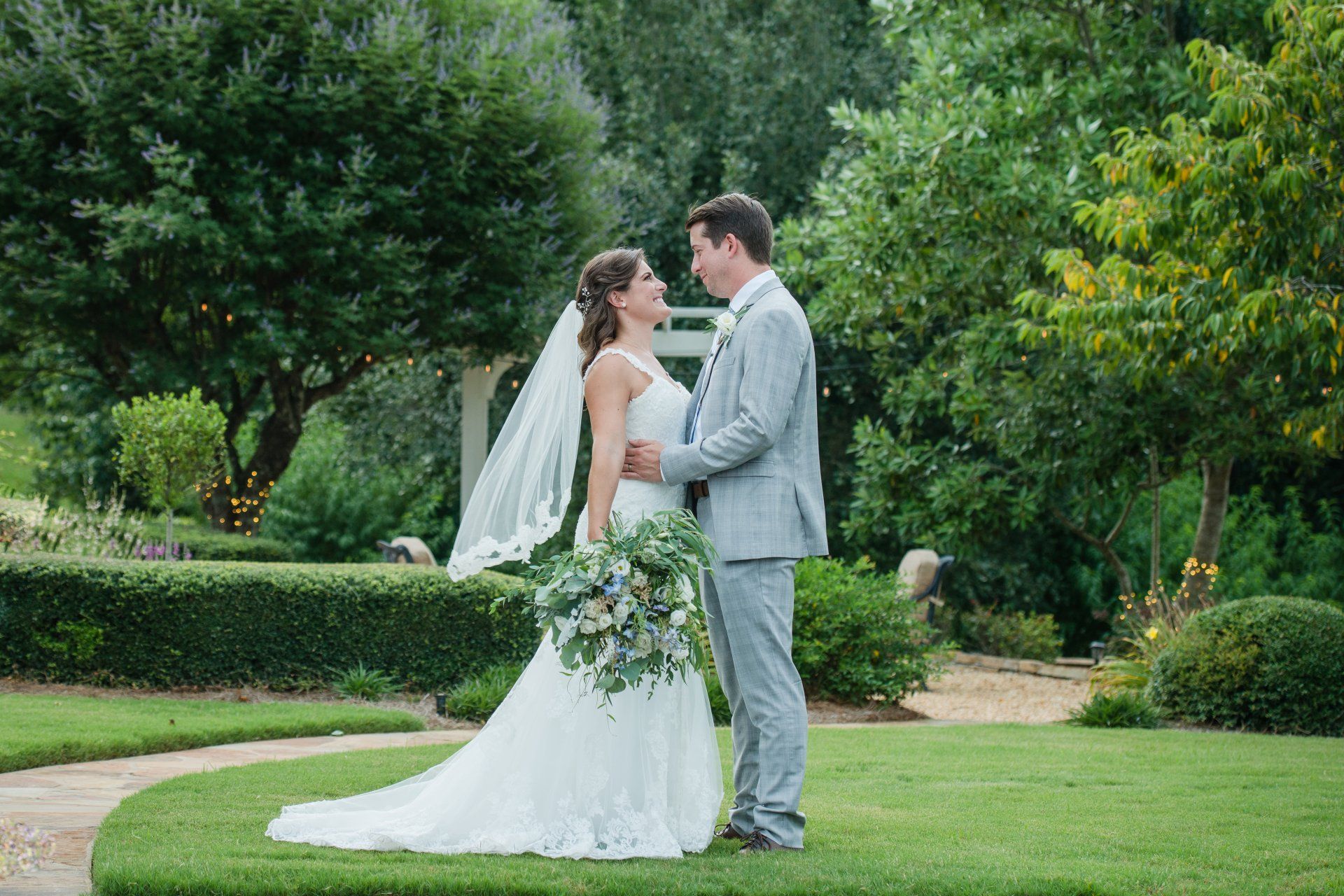 Bride and groom in white and gray wedding attire standing in a green garden, facing each other.