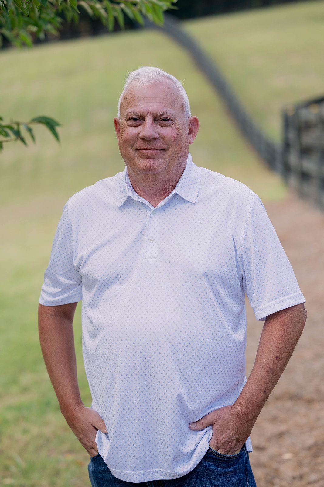 Smiling man in a white shirt standing outdoors on a grassy path with hands in pockets