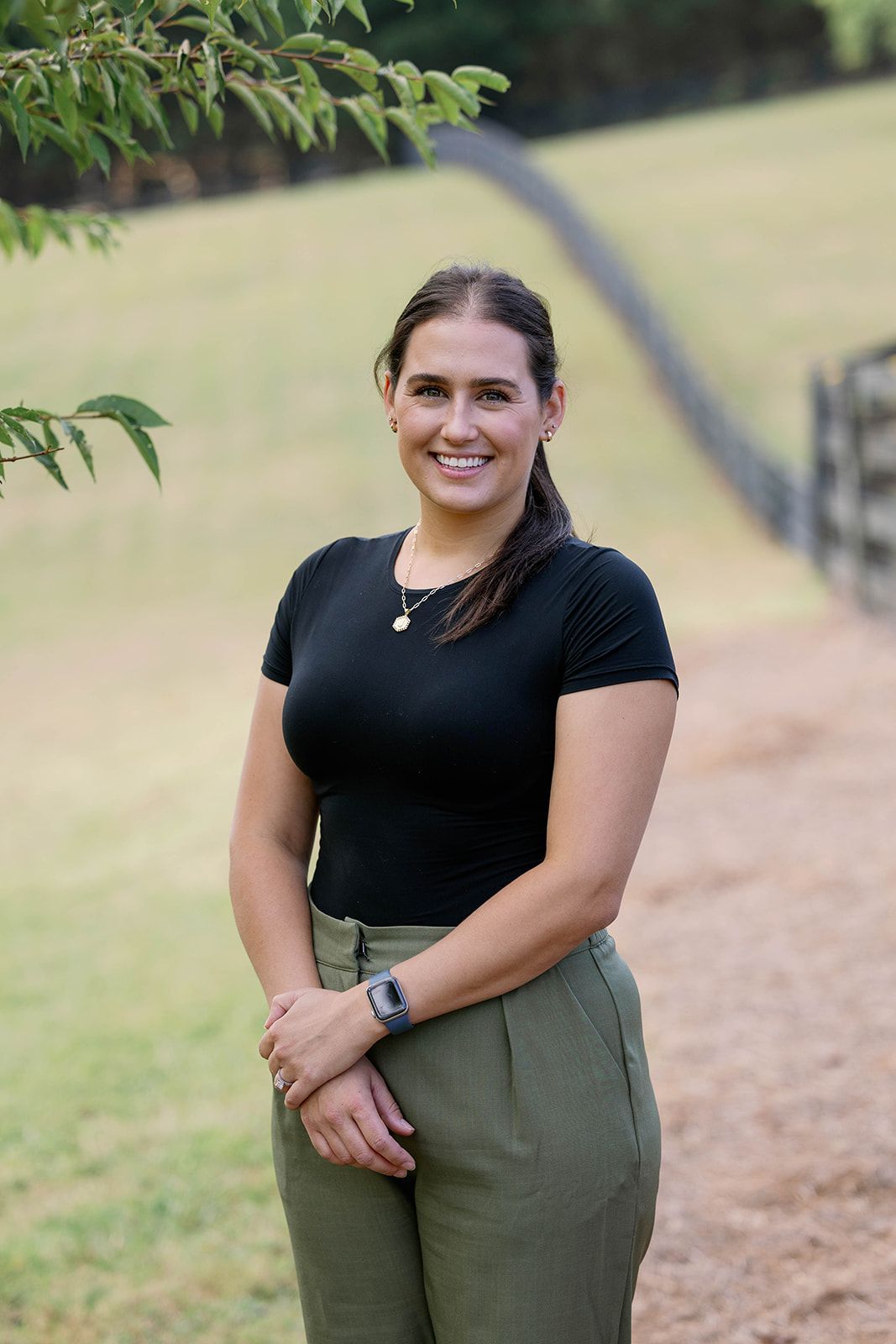 Person standing outdoors on a path, wearing a black top and olive pants, smiling at the camera.