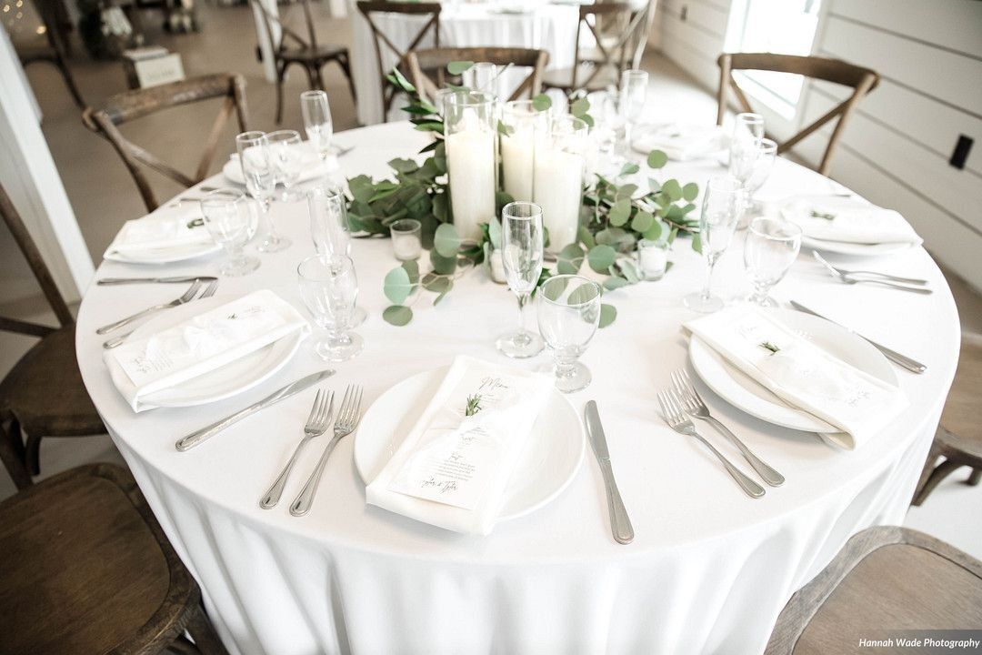 Round white banquet table set with white napkins, silverware, candles, and greenery centerpiece