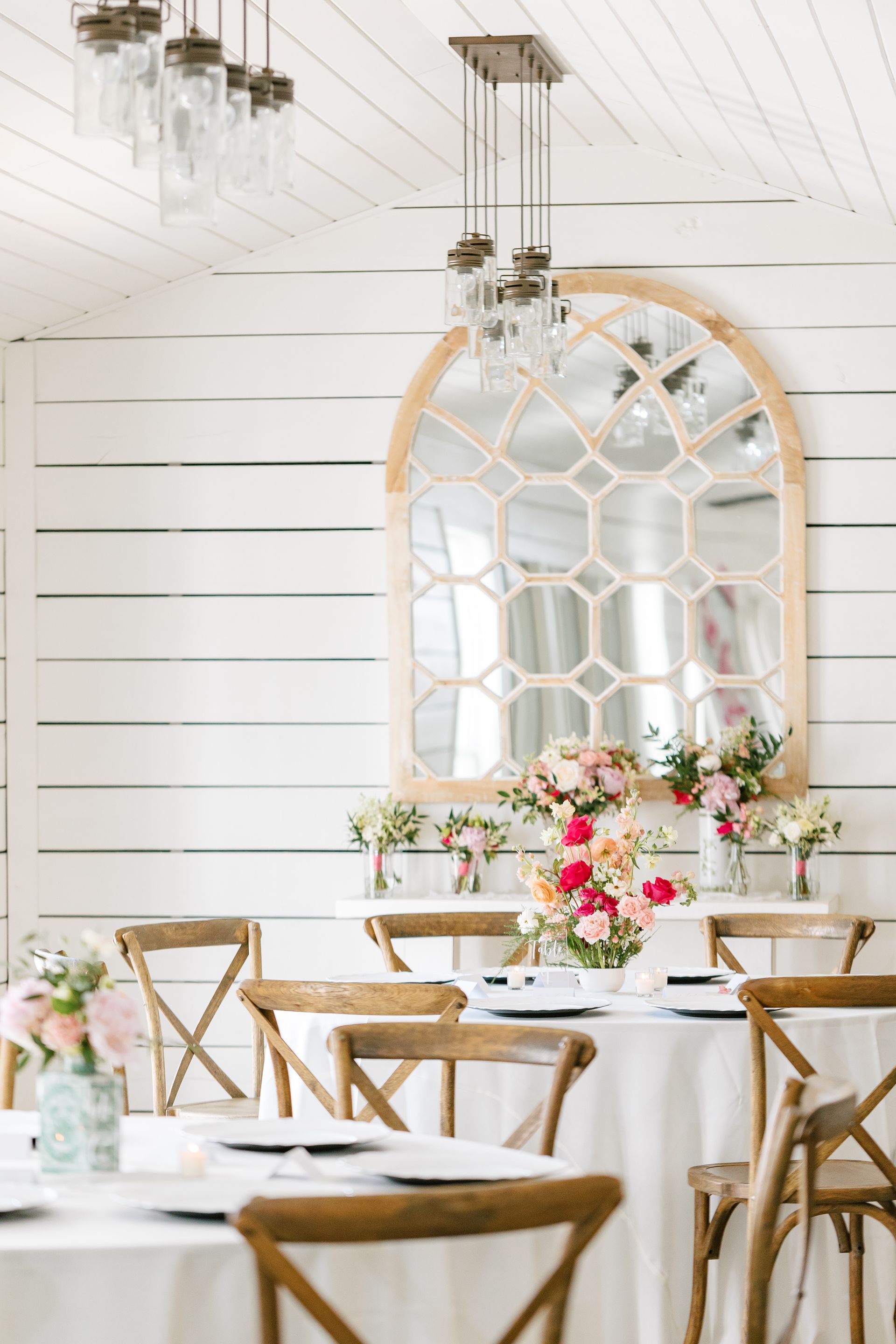Elegant white dining setup with floral centerpieces, gold chairs, and a large arched mirror on a bright wall.