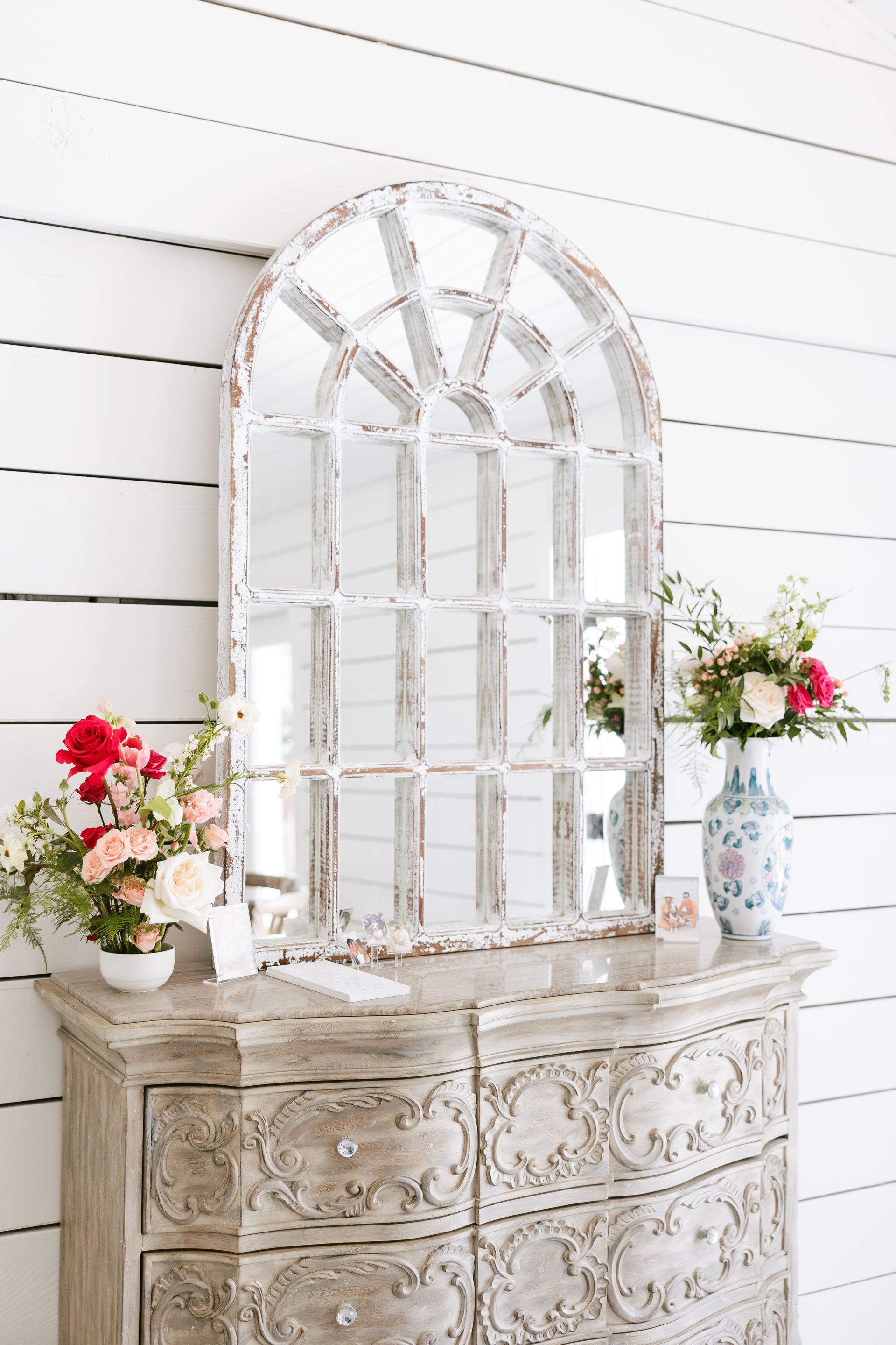White arched mirror on ornate dresser with floral arrangements against a white shiplap wall
