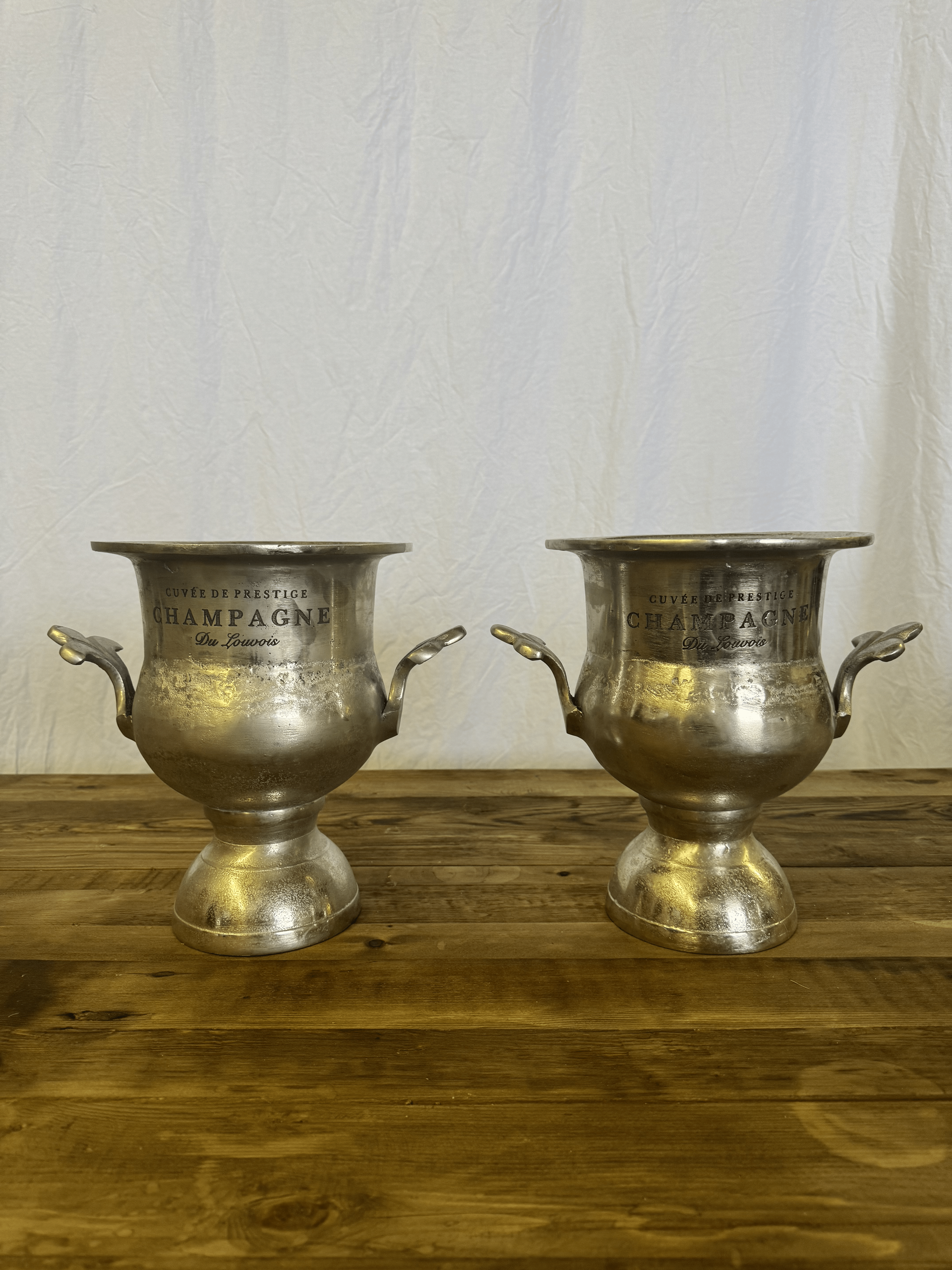 Two silver trophy cups with handles sit on a wooden table against a plain white wall.