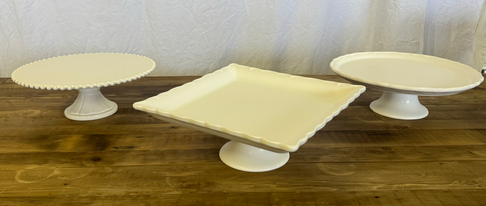 White pedestal cake stands and a square platter on a wooden table