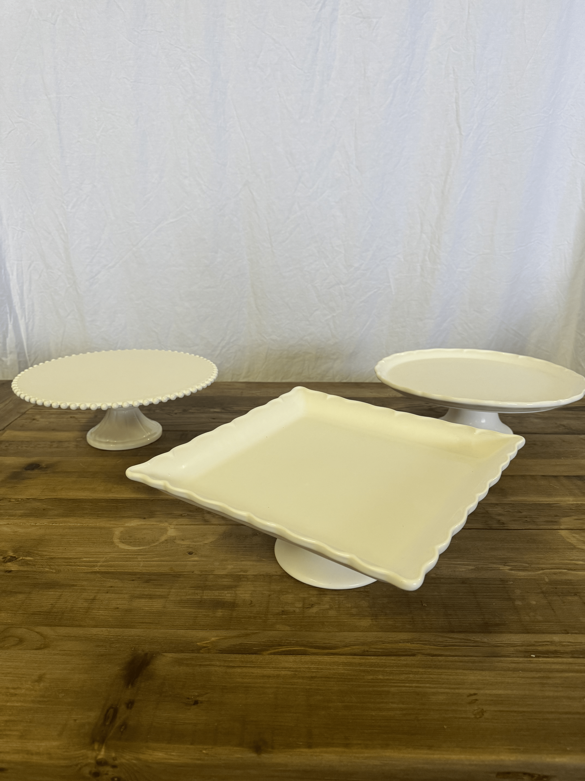 White cake stands with a square tray on a wooden table against a plain wall