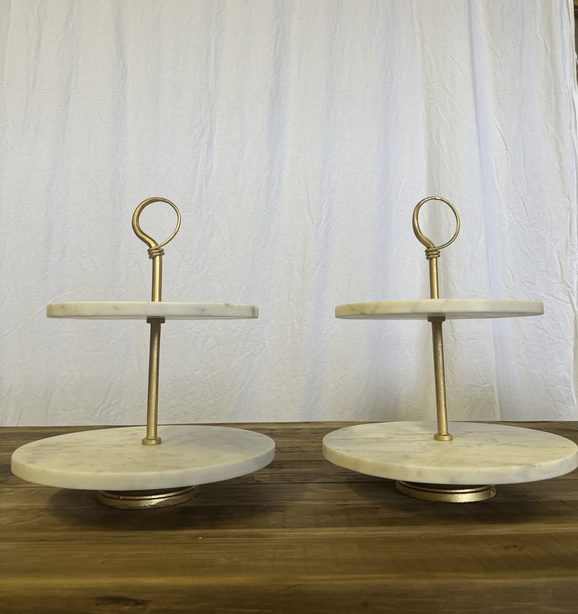 Two beige two-tier serving trays with gold handles on a wooden table against a white curtain backdrop