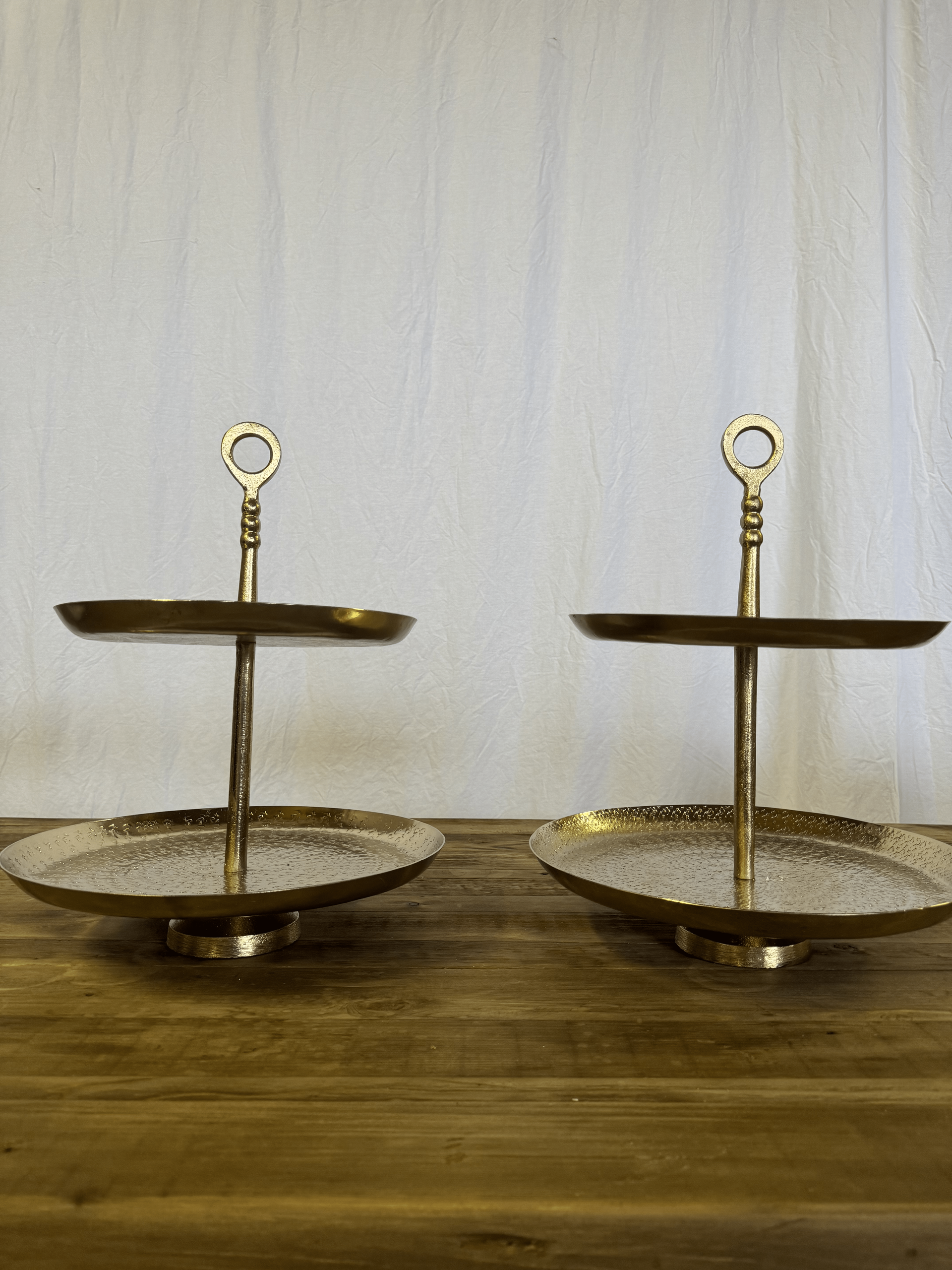Pair of gold-toned tiered serving stands on a wooden table against a white backdrop