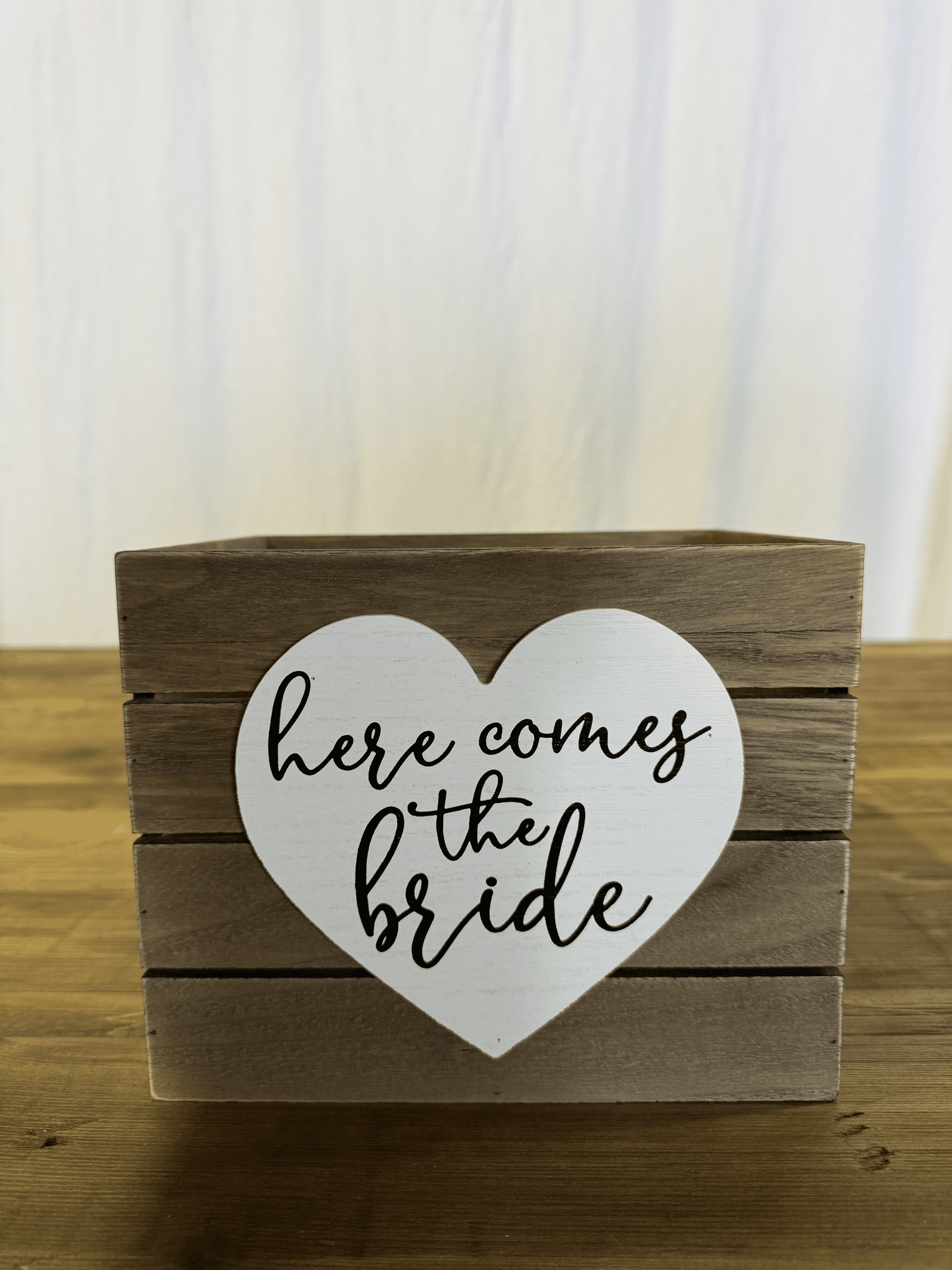 Wooden block with white heart reading “here comes the bride” on a wood surface