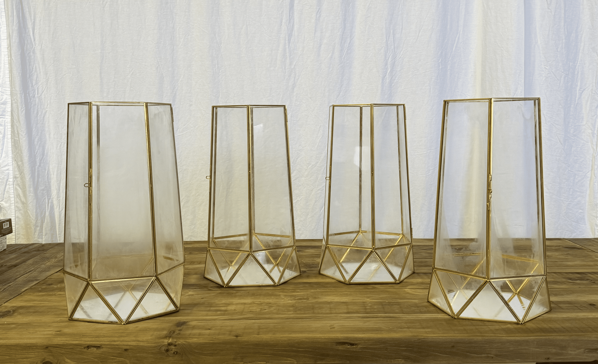Four gold geometric candle holders on a wooden surface against a white curtain.