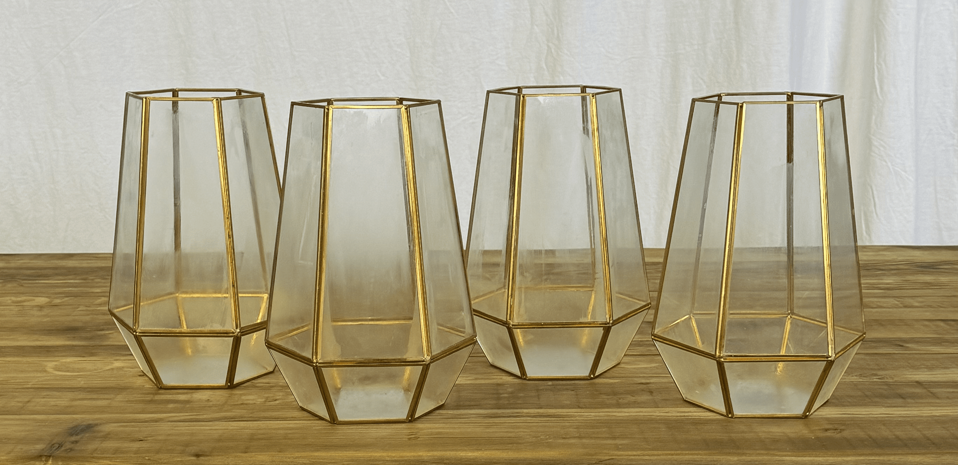Four gold geometric glass candle holders on a wooden surface with a light curtain backdrop