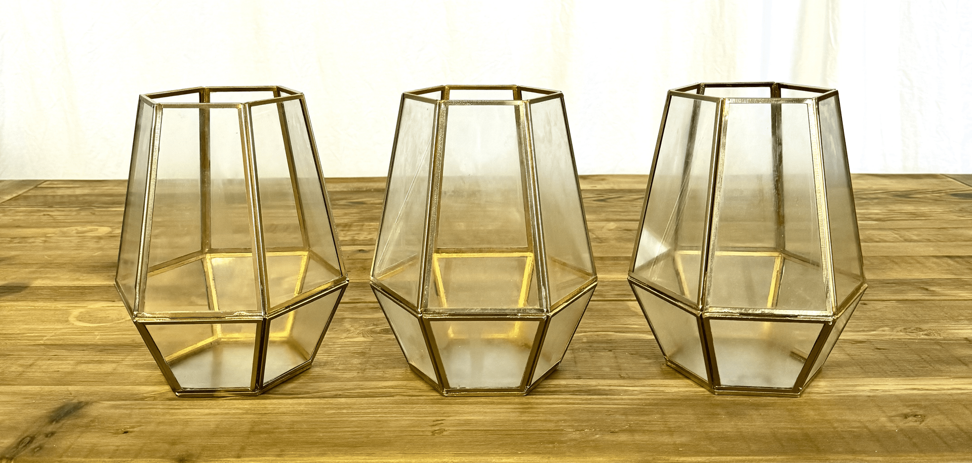 Three gold geometric glass candle holders on a wooden table against a white wall