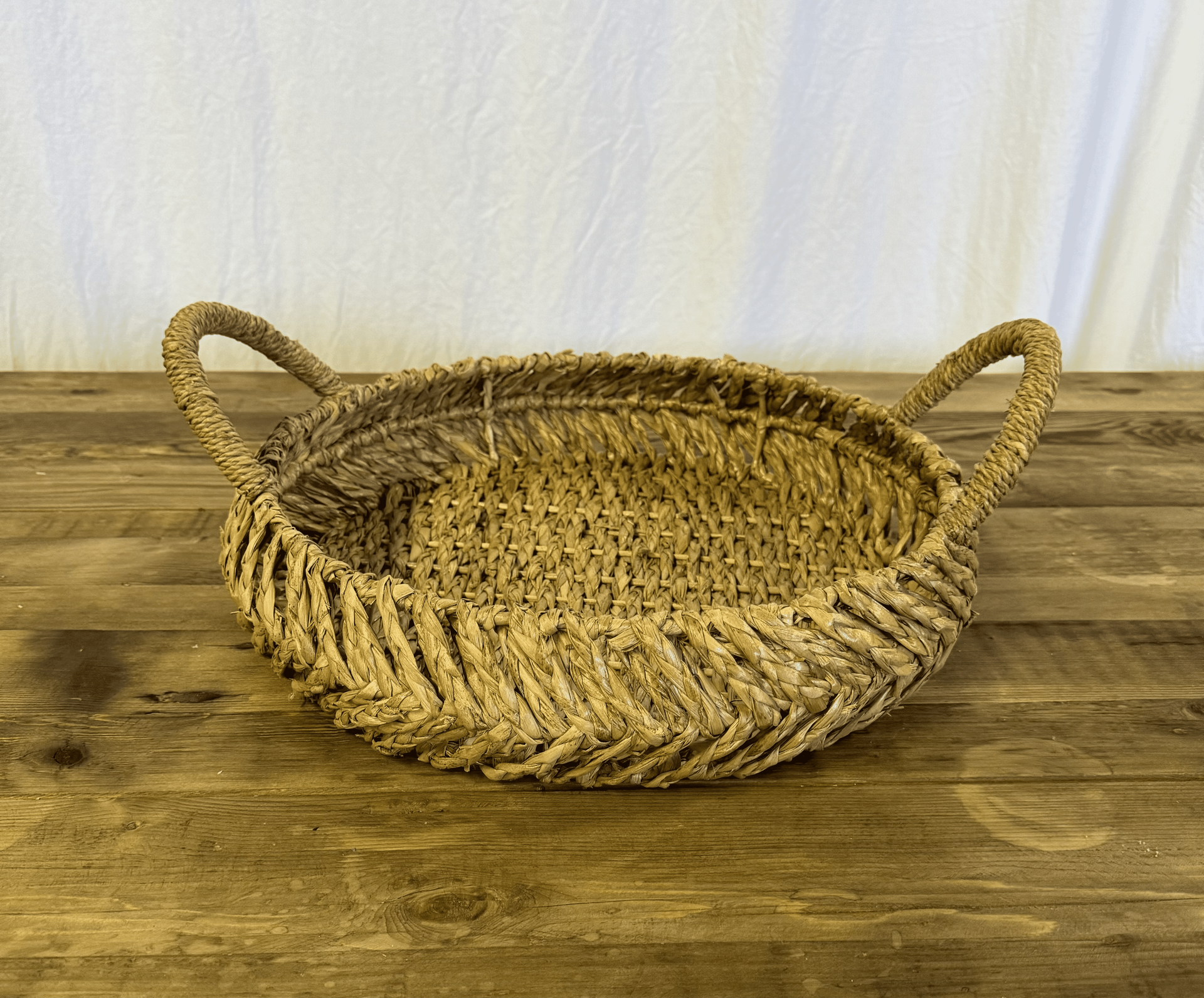 Oval woven straw basket with two handles on a wooden table against a light curtain background