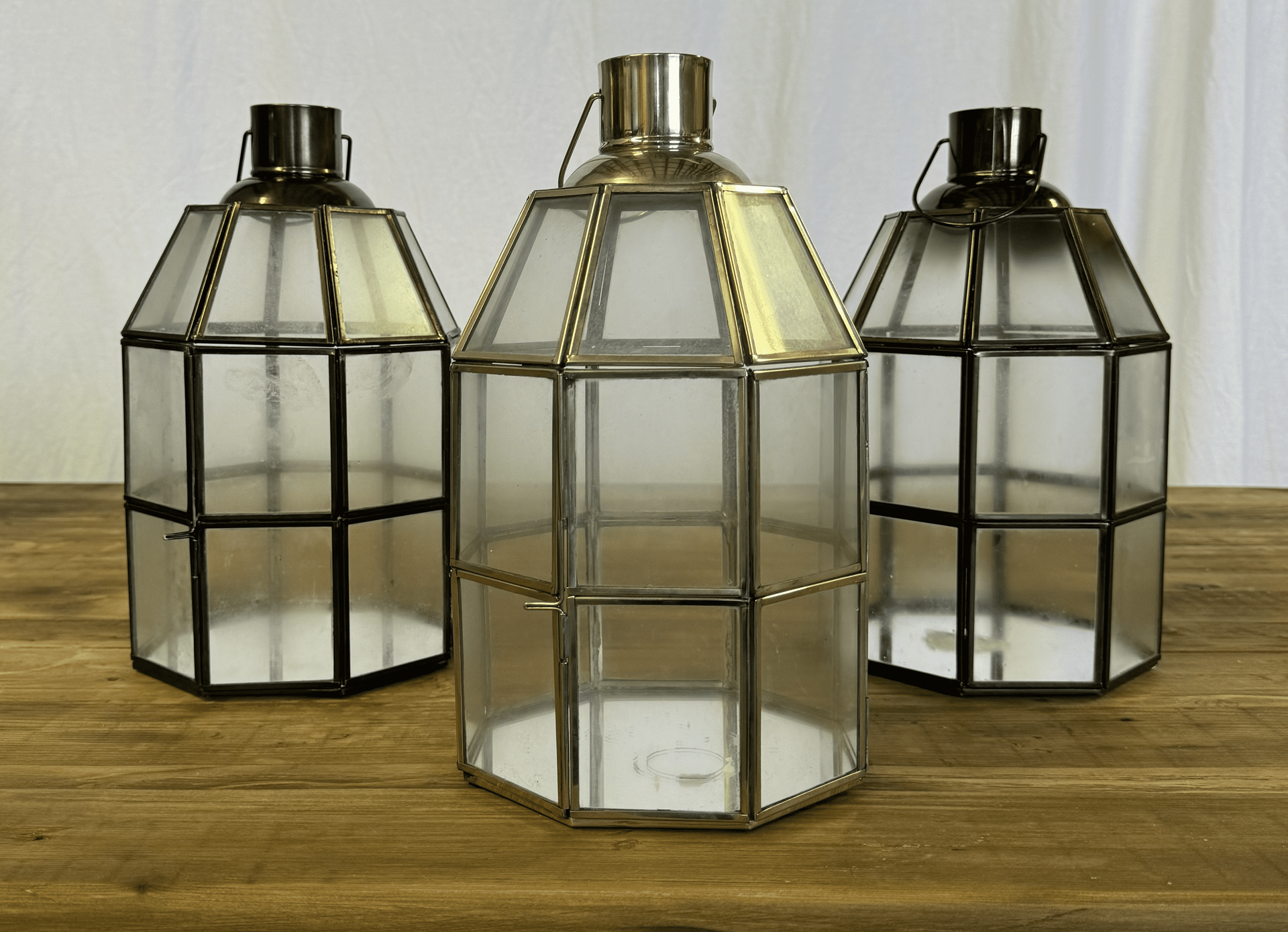 Three vintage lantern-style candle holders on a wooden table, with clear and black metal frames.