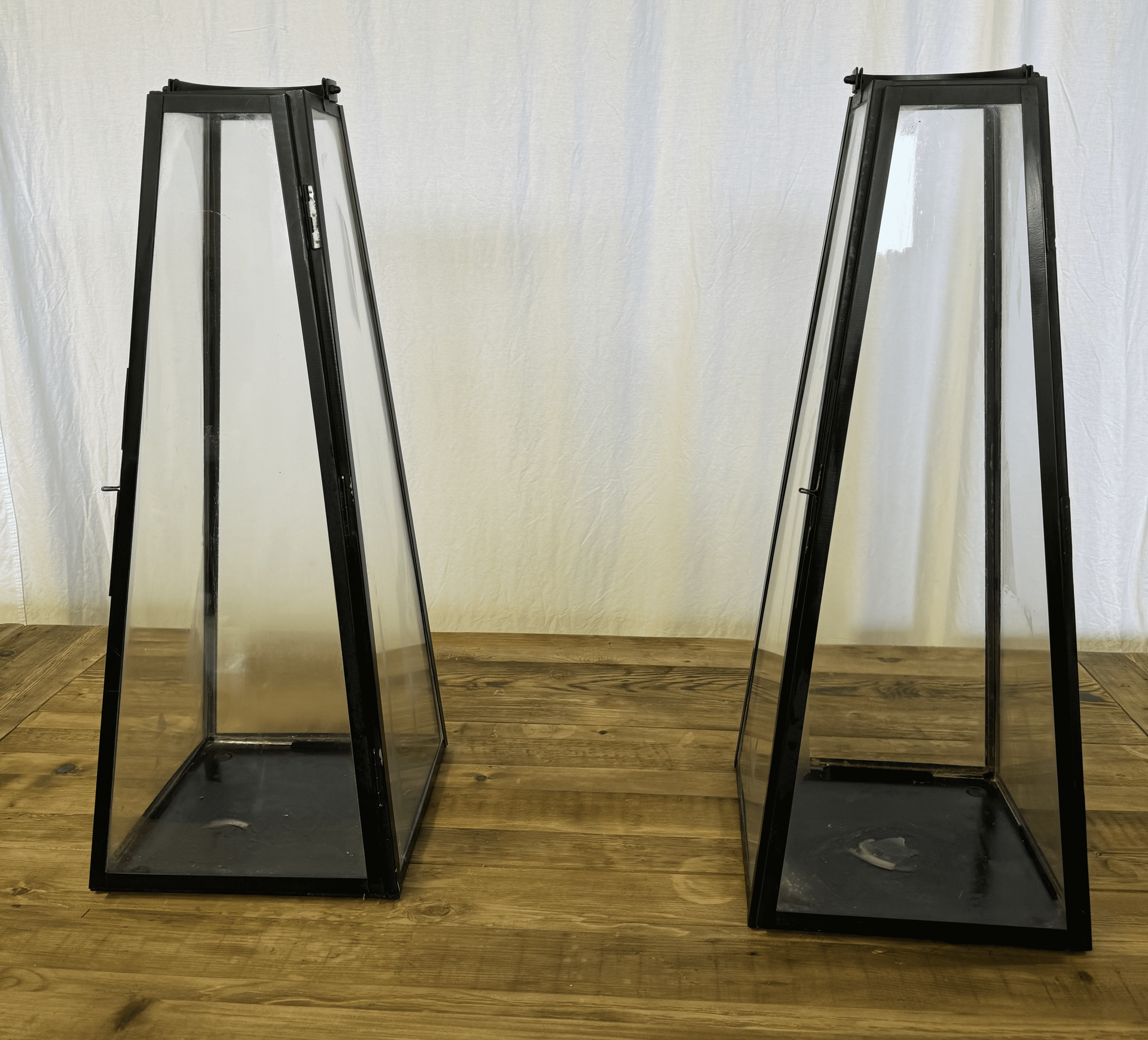 Two black tapered metal plant stands on a wooden floor against a light curtain backdrop