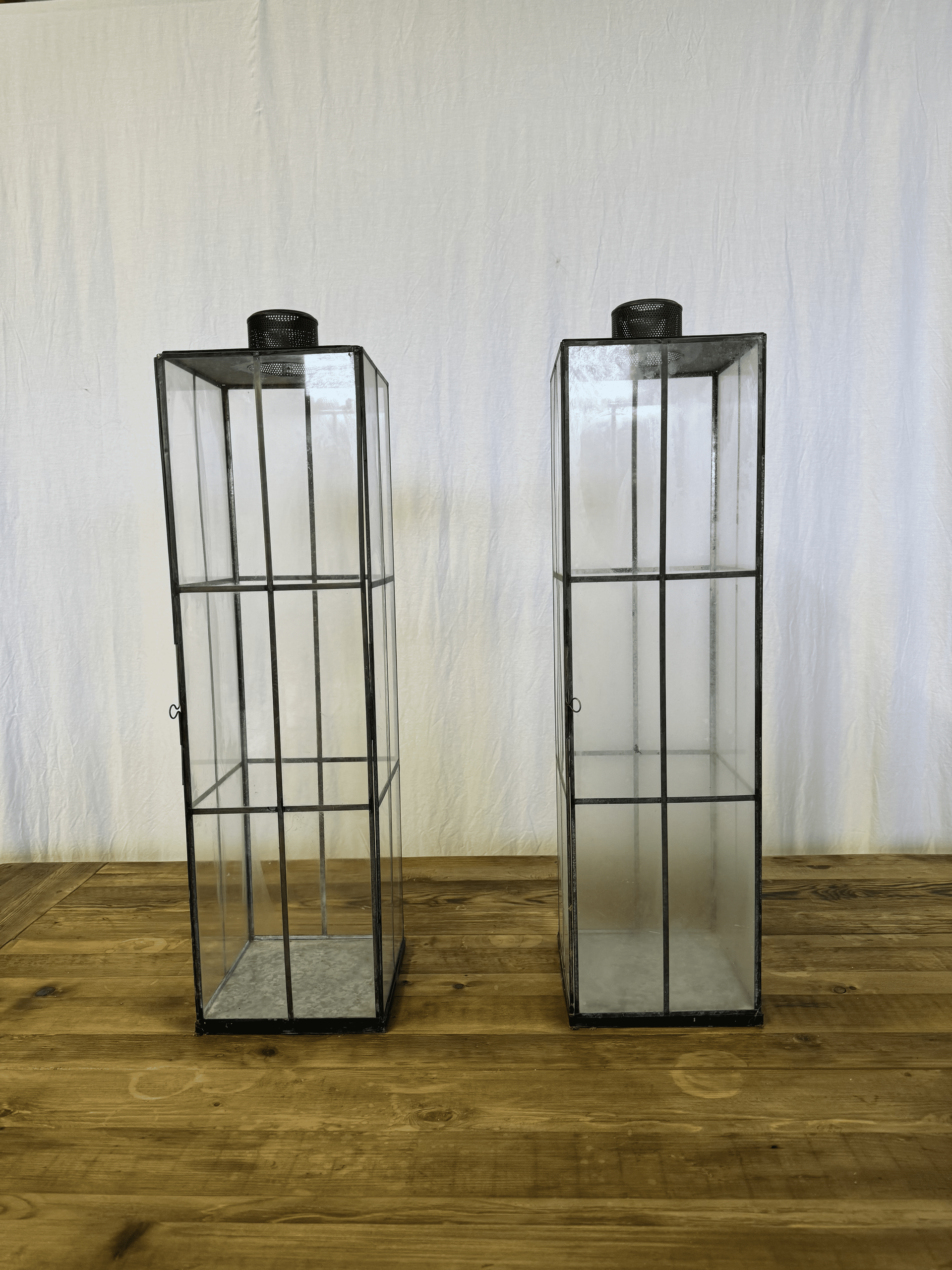 Two tall wire cage lanterns on a wooden surface against a plain wall.