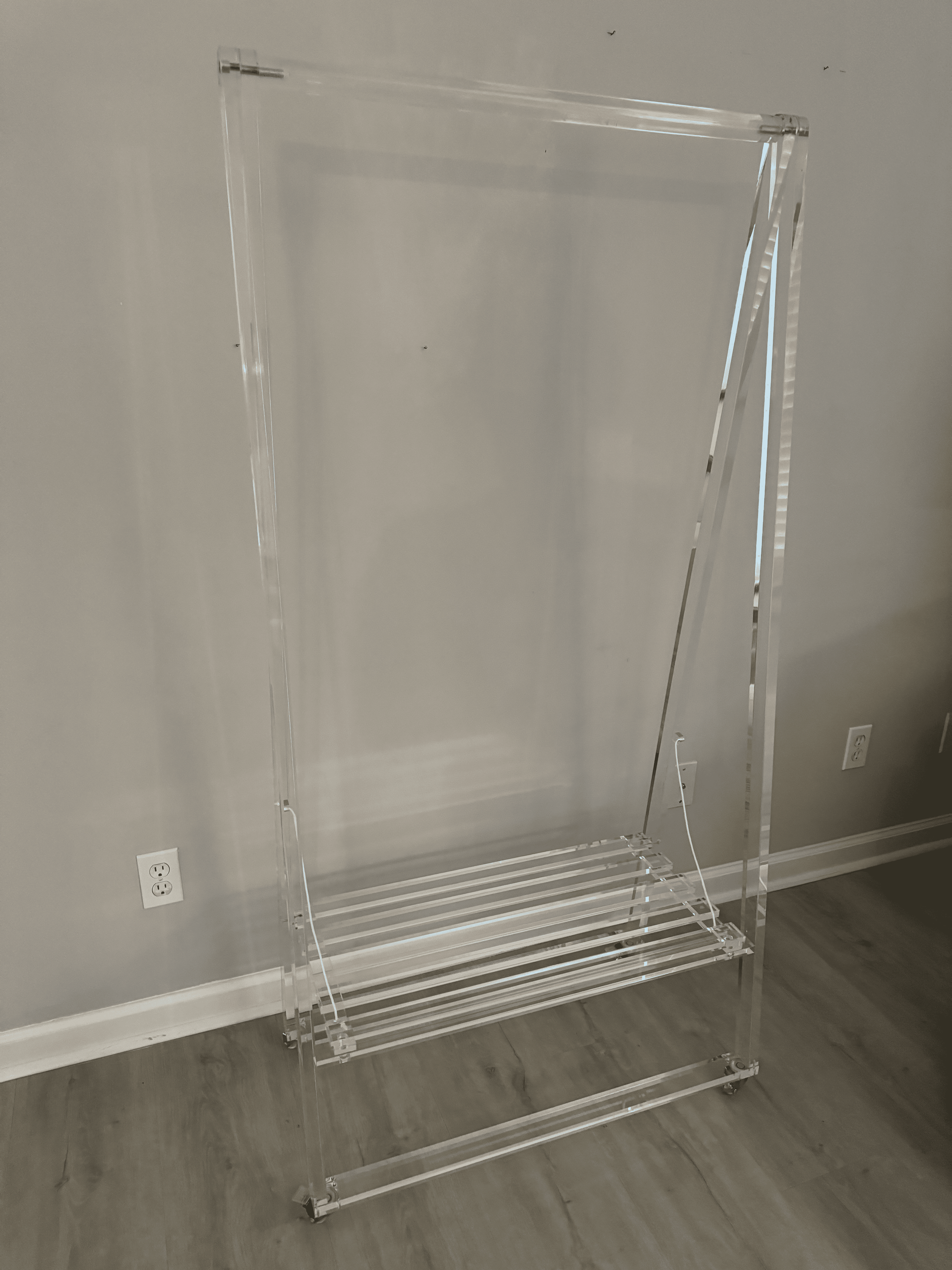 Clear acrylic garment rack frame against a white wall in a room with hardwood floor.