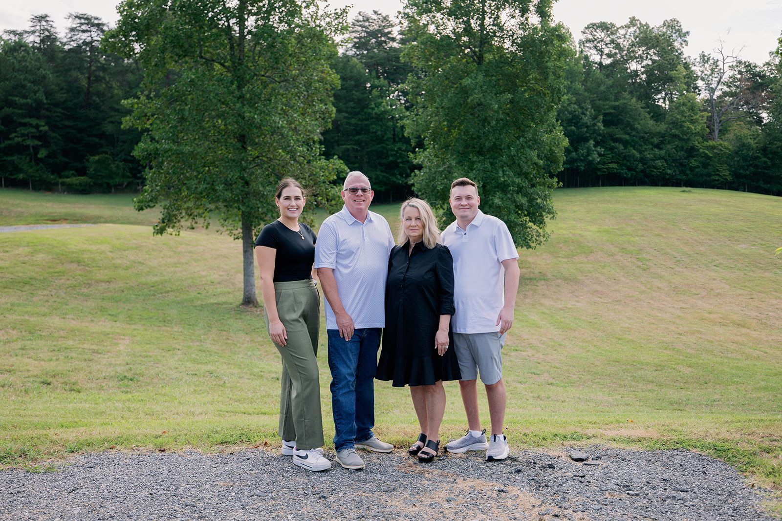 Four people standing on gravel in a grassy park, with trees and a small hill behind them.