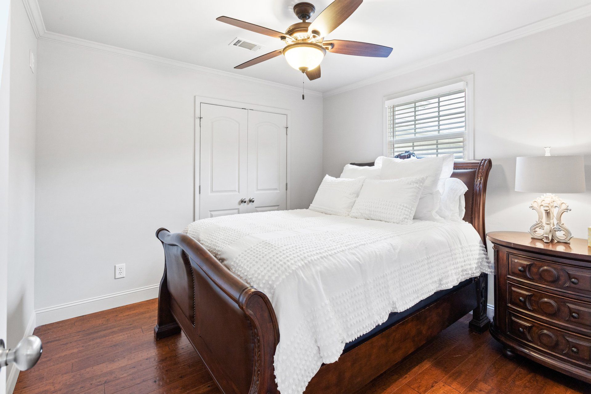 Bright bedroom with dark wood bed, white bedding, hardwood floors, and ceiling fan