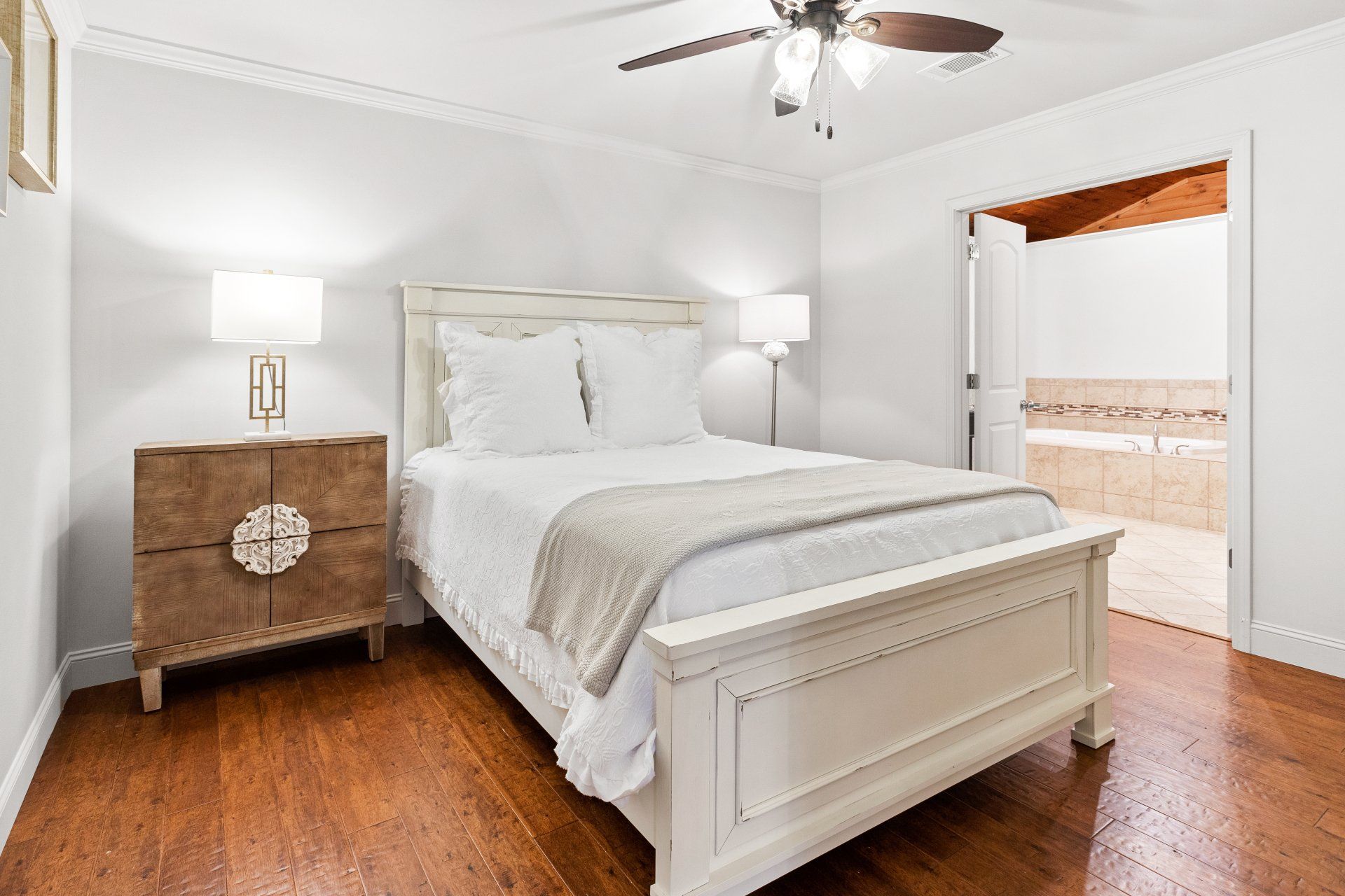 Bright bedroom with white bed, wood floor, nightstands, ceiling fan, and open doorway to bathroom