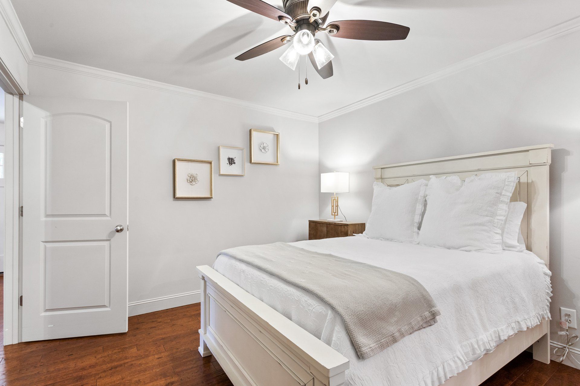 Bright white bedroom with a bed, ceiling fan, framed art, and wooden floor