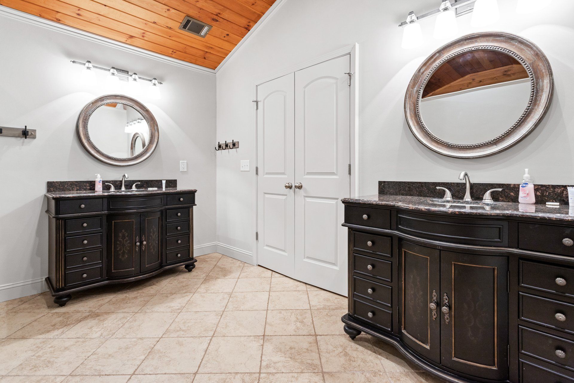 Elegant bathroom with dark wood vanities, round mirrors, white walls, and a wood-paneled ceiling